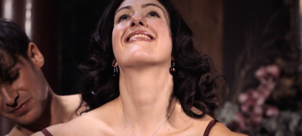 Sadie Lune smiles happily while Parker Marx dresses her in a bra in Adorn, an erotic game film directed by Jennifer Lyon Bell for Blue Artichoke Films